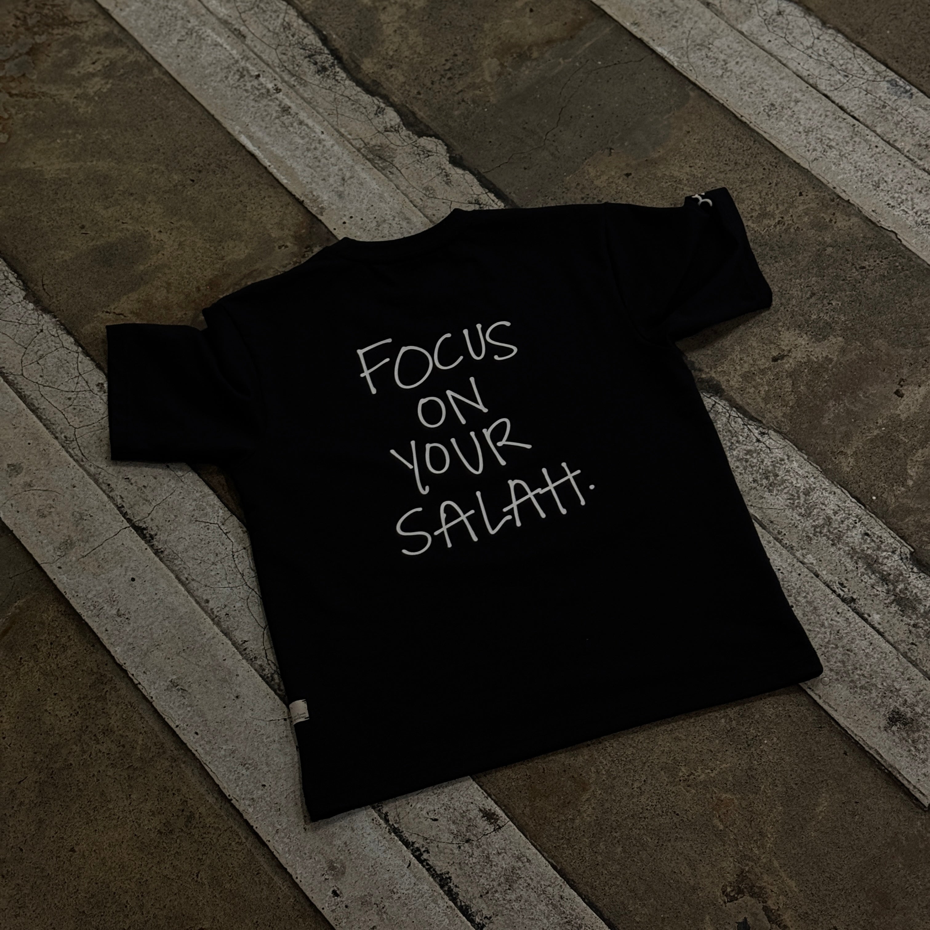 Black t-shirt with 'Focus on Your Salah' text on a stone floor.