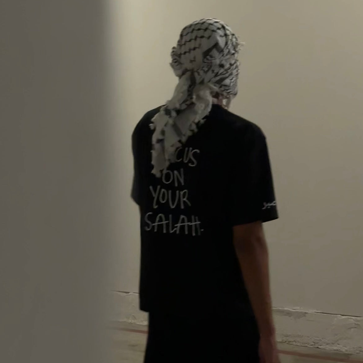 Person wearing a black t-shirt with text and a headscarf in a neutral setting