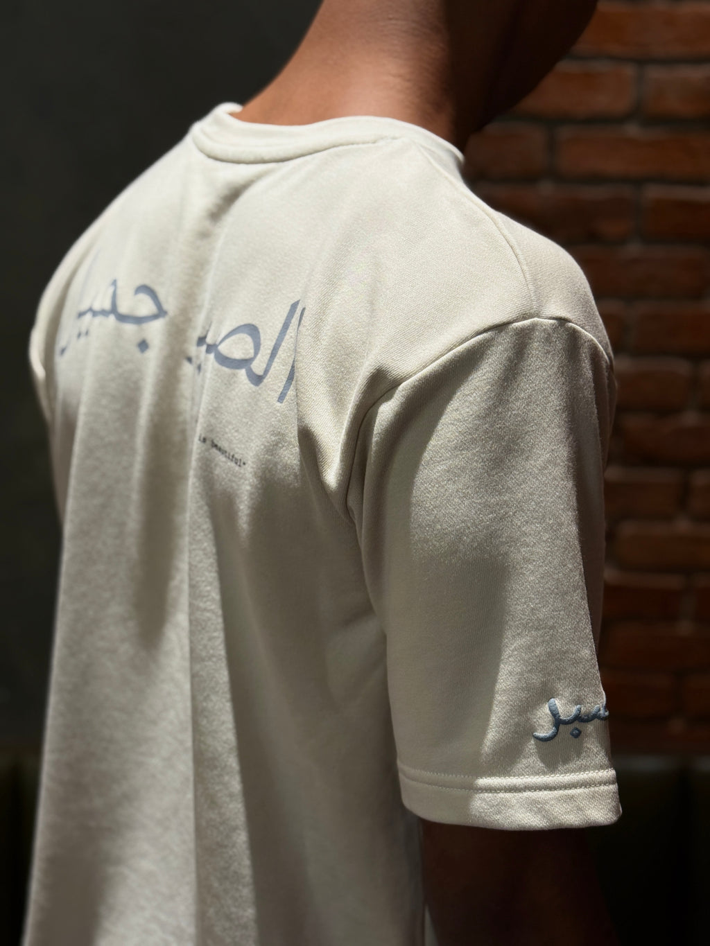 Person wearing a light-colored t-shirt with Arabic text on the back, standing against a brick wall.