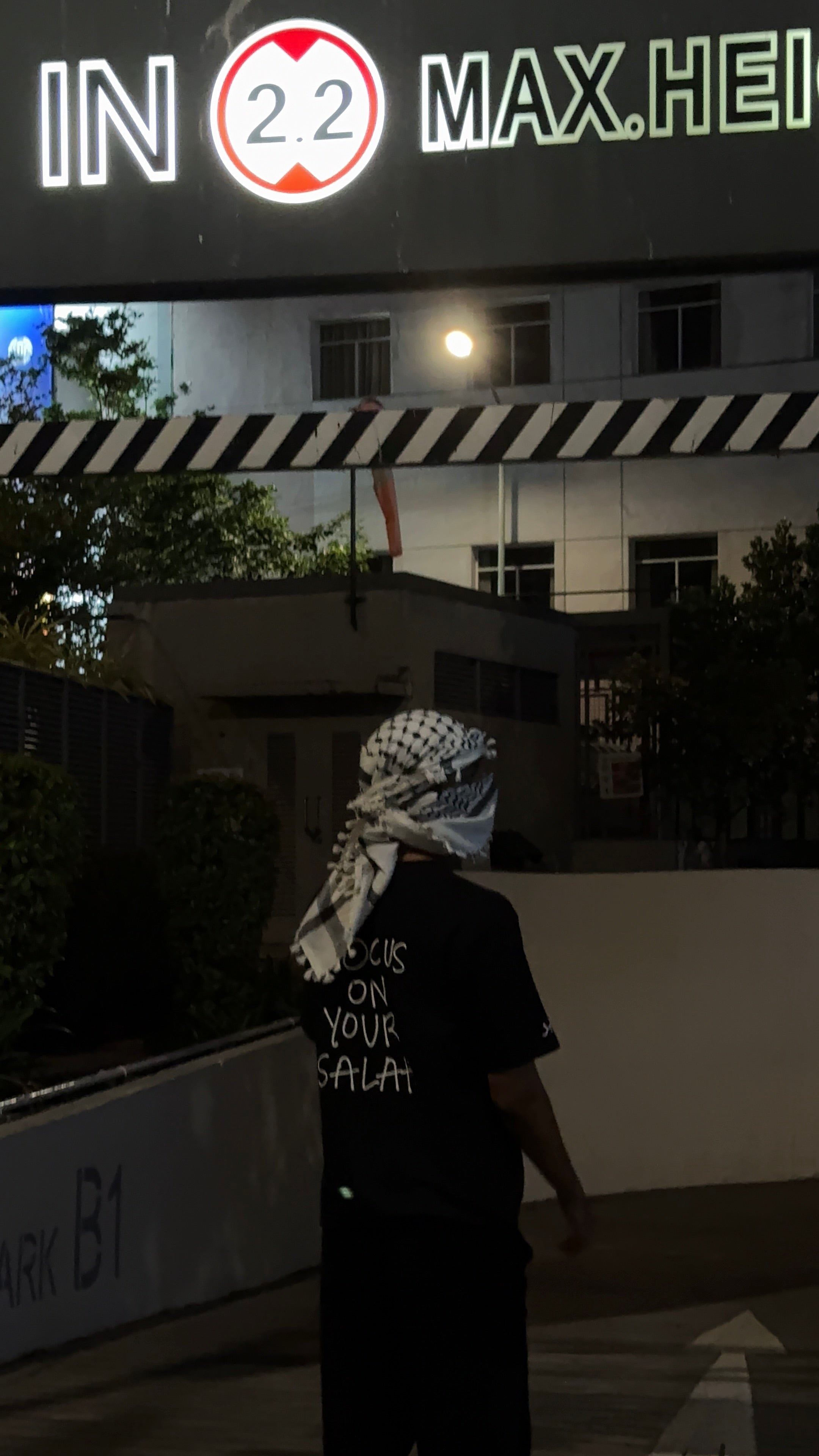 Person wearing a checkered headscarf walking under a neon sign at night.