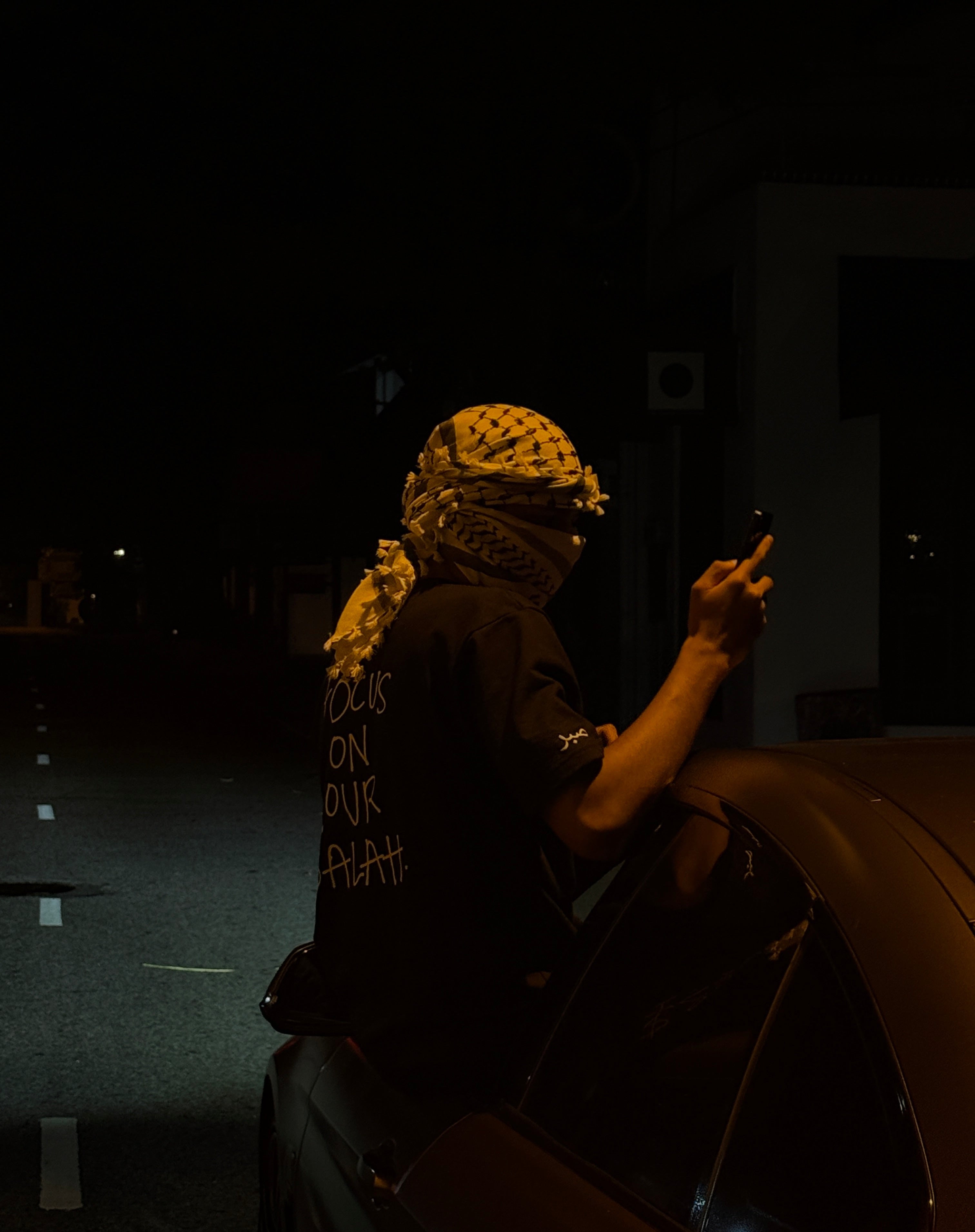 Person sitting on a car at night, wearing a headscarf and a shirt with text.