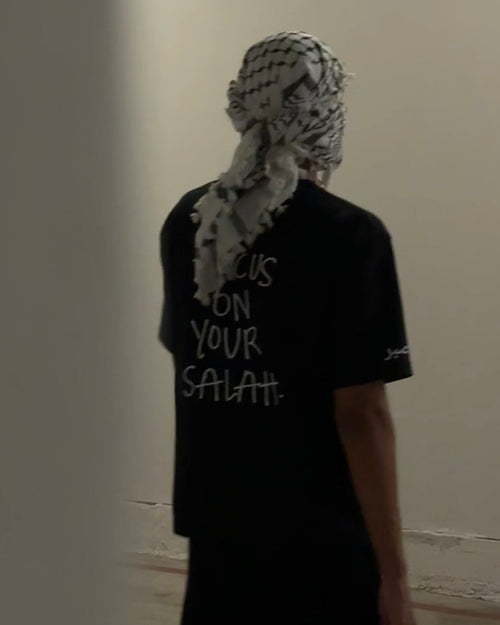 Person wearing a black t-shirt with text and a headscarf in a neutral setting